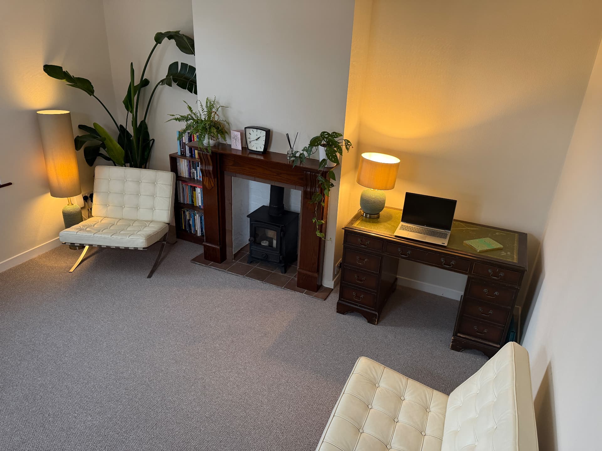 The therapy room in warm evening light — leather chairs by the wood-burner, bookshelf and a writing desk